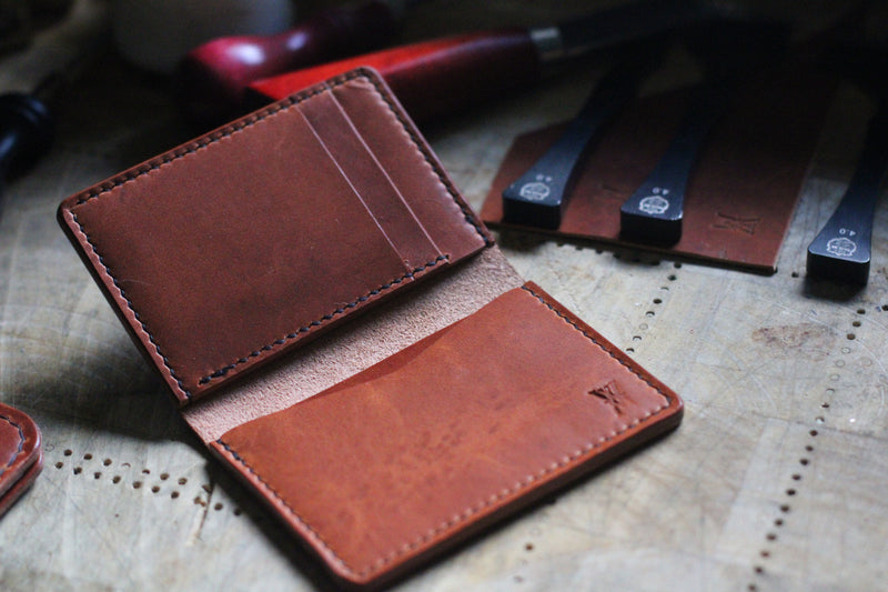 A brown Horween Dublin leather card case from the WM Leatherwork Collection by Rogue Industries is open on a wooden surface with various leather tools in the background, showcasing the beauty of a handcrafted leather piece.