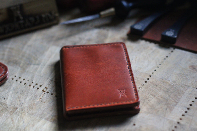 A WM Leatherwork Collection - Premium Leather Card Case by Rogue Industries, crafted from Horween Dublin leather, rests closed on a wooden surface, surrounded by leatherworking tools.