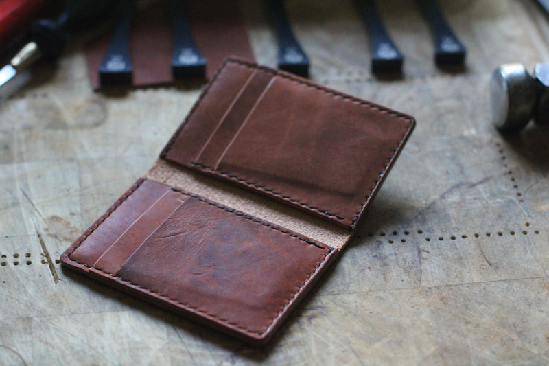 A **WM Leatherwork Collection - Premium Leather Card Case** from **Rogue Industries**, made of hand-crafted brown Horween Dublin leather, is open on a wooden surface, displaying its minimalist card slots with leatherworking tools in the background.