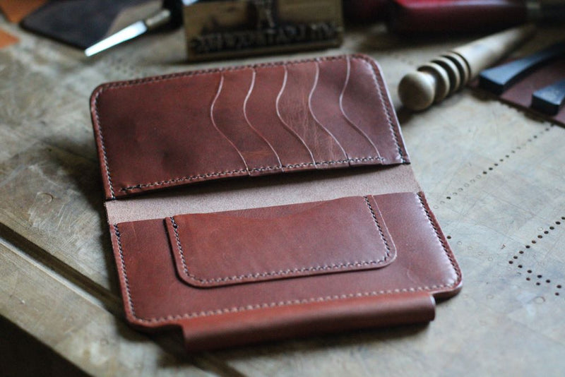 The WM Leatherwork Collection - Premium Leather Field Notes Wallet from Rogue Industries lies open on a wooden surface, revealing its multiple card slots and pocket. Various leatherworking tools are scattered in the background.