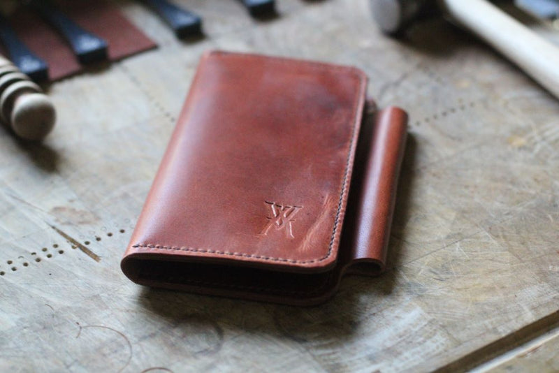 A brown leather Rogue Industries Wallet from the WM Leatherwork Collection, featuring a fold-over design and stamped logo, lies on a wooden surface surrounded by various tools.