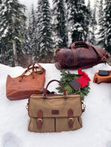 The Rogue Industries Waxed Canvas Laptop Bag, crafted from waxed canvas and top grain leather, alongside a wallet, is artfully displayed on snow near evergreen trees, accented by a red bow decoration.