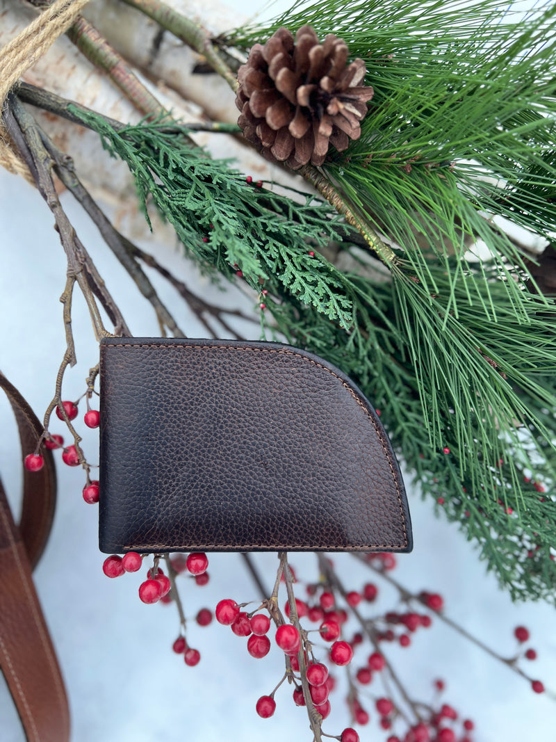 A Rogue Industries Classic Made in Maine Edition Front Pocket Wallet, featuring luxurious brown leather craftsmanship, is elegantly displayed on a pine branch decorated with a pine cone and bright red berries.